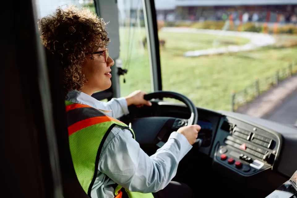Woman bus driver sitting behind wheel of bus wearing long-sleeved shirt and yellow safety vest.